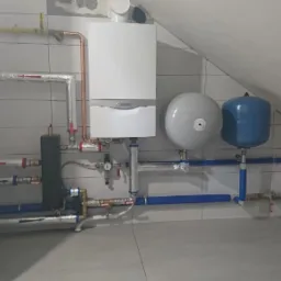 Biały piec gazowy Vaillant z miedzianymi rurami, szare i niebieskie zbiorniki wyrównawcze oraz niebieskie rury instalacji w pomieszczeniu wyłożonym płytkami.