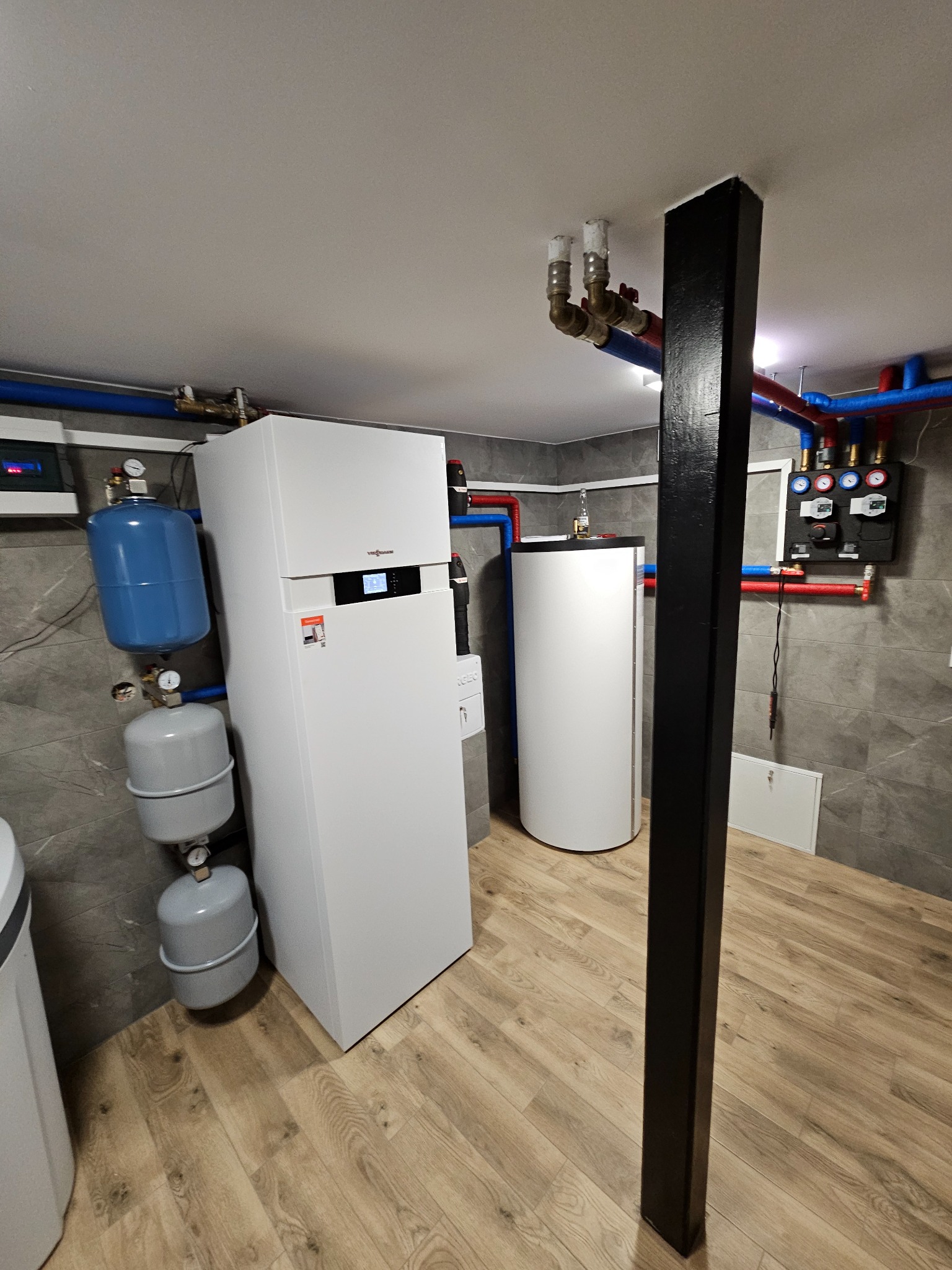 Nowoczesna kotłownia: biały piec Viessmann, zbiornik buforowy, naczynia przeponowe, instalacja w rurach PEX, szara płytka na ścianie, drewnopodobna podłoga.