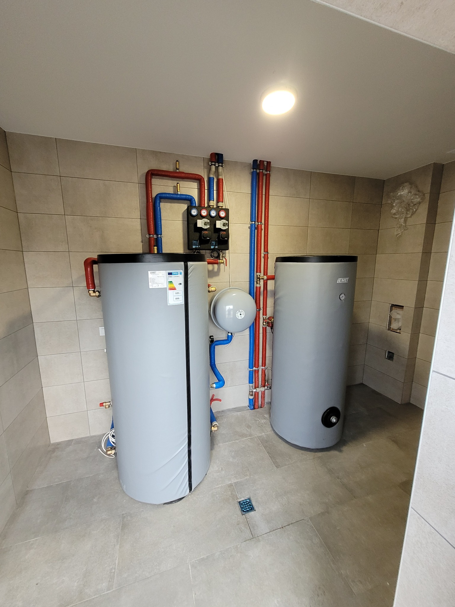 Dwa szare zbiorniki ciepłej wody użytkowej z podłączonymi rurami (czerwone i niebieskie) w kotłowni wykończonej płytkami ceramicznymi. Widoczne zawory i manometry.