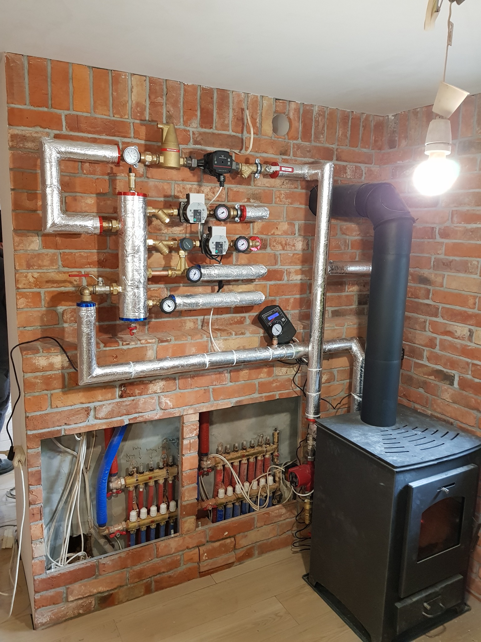 Instalacja CO z piecem na drewno na tle ceglanej ściany. Rury z izolacją aluminiową, zawory, manometry. Rozdzielacz ogrzewania podłogowego w dolnej części.