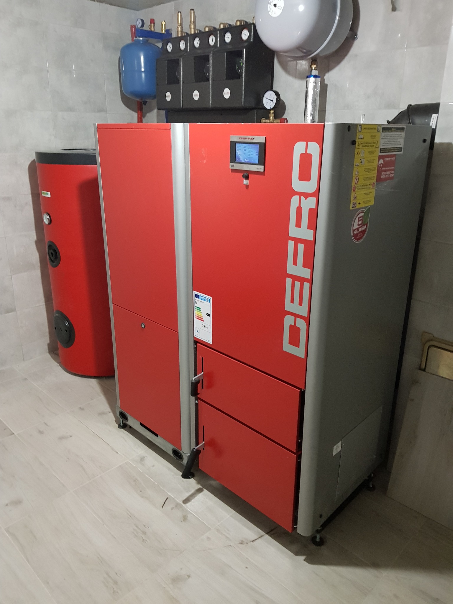 Czerwono-szary piec CO marki Defro w kotłowni z zbiornikiem na wodę. Widoczne elementy instalacji, manometry i zabezpieczenia. Kotłownia wykończona płytkami.