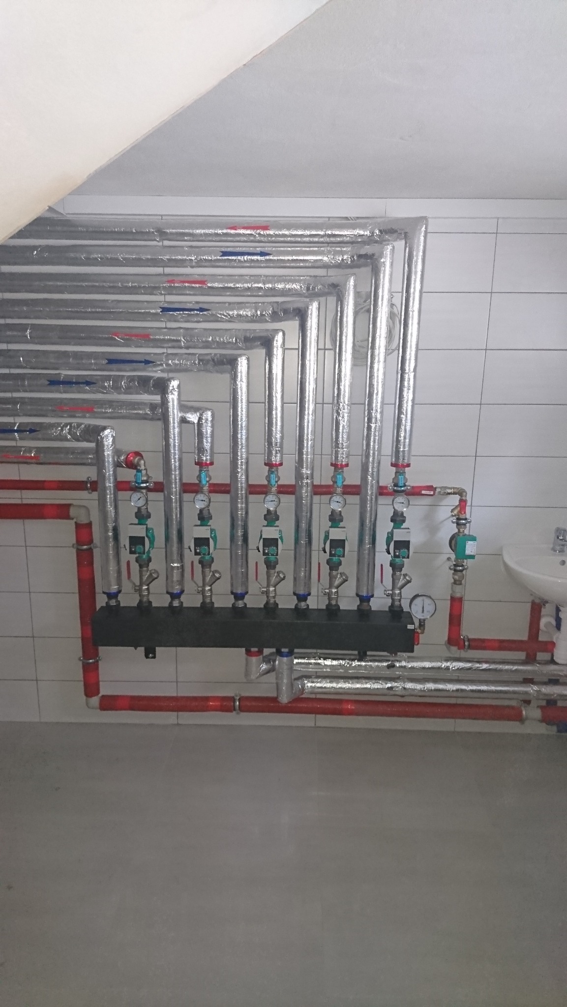 Rozbudowana instalacja grzewcza z licznymi rurami, zaworami i manometrami, częściowo zaizolowana, zamontowana przy ścianie z białych płytek w pomieszczeniu.