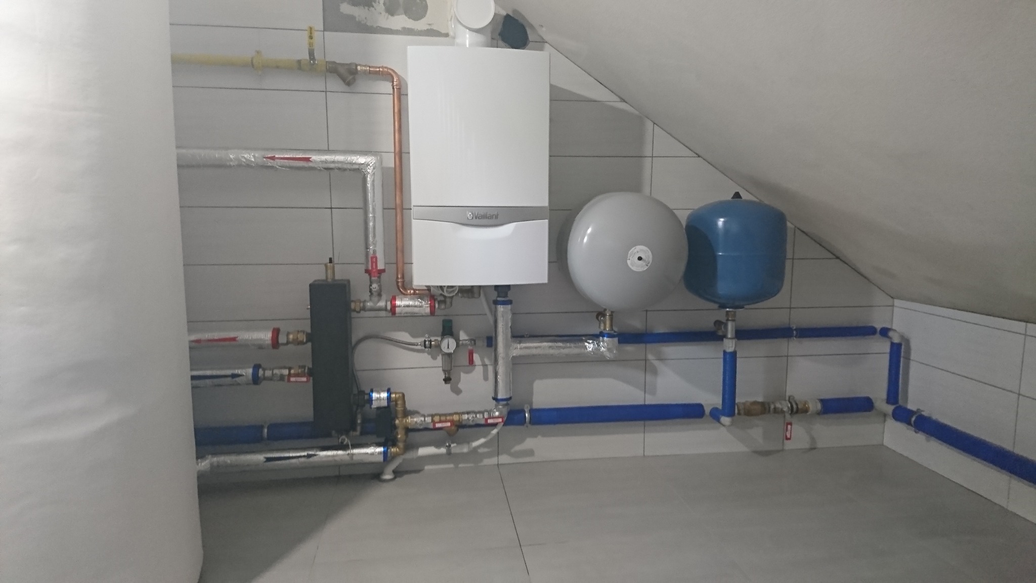 Biały piec gazowy Vaillant z miedzianymi rurami, szare i niebieskie zbiorniki wyrównawcze oraz niebieskie rury instalacji w pomieszczeniu wyłożonym płytkami.