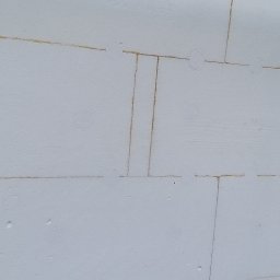 WORK-JAW - Białe płyty styropianowe z widocznymi łączeniami, tworzące izolację ściany zewnętrznej budynku w trakcie budowy. Fragment rusztowania po prawej stronie.