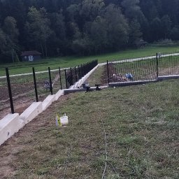 WORK-JAW - Montaż ogrodzenia panelowego na betonowej podmurówce w terenie zielonym. Widoczne narzędzia i materiały budowlane. W tle las i mały budynek.