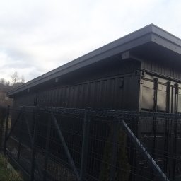 WORK-JAW - Czarna wiata z kontenerów morskich, otoczona metalowym ogrodzeniem panelowym na tle jesiennego krajobrazu. Widoczne detale konstrukcyjne i brama wjazdowa.