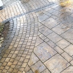 WORK-JAW - Czyszczenie kostki brukowej z użyciem myjki ciśnieniowej. Widoczne mokre i czyste fragmenty kostki, co sugeruje etap pracy w toku. Tło: trawnik i elementy dekoracyjne.