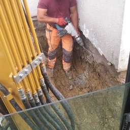 WORK-JAW - Pracownik w kasku i roboczych spodniach używa młota pneumatycznego przy fundamencie budynku. Widoczny fragment koparki z żółtymi przewodami.