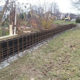 WORK-JAW - Zbrojenie fundamentu pod ogrodzenie z izolacją pionową, widoczny szalunek i wykop. W tle domy i koparka.