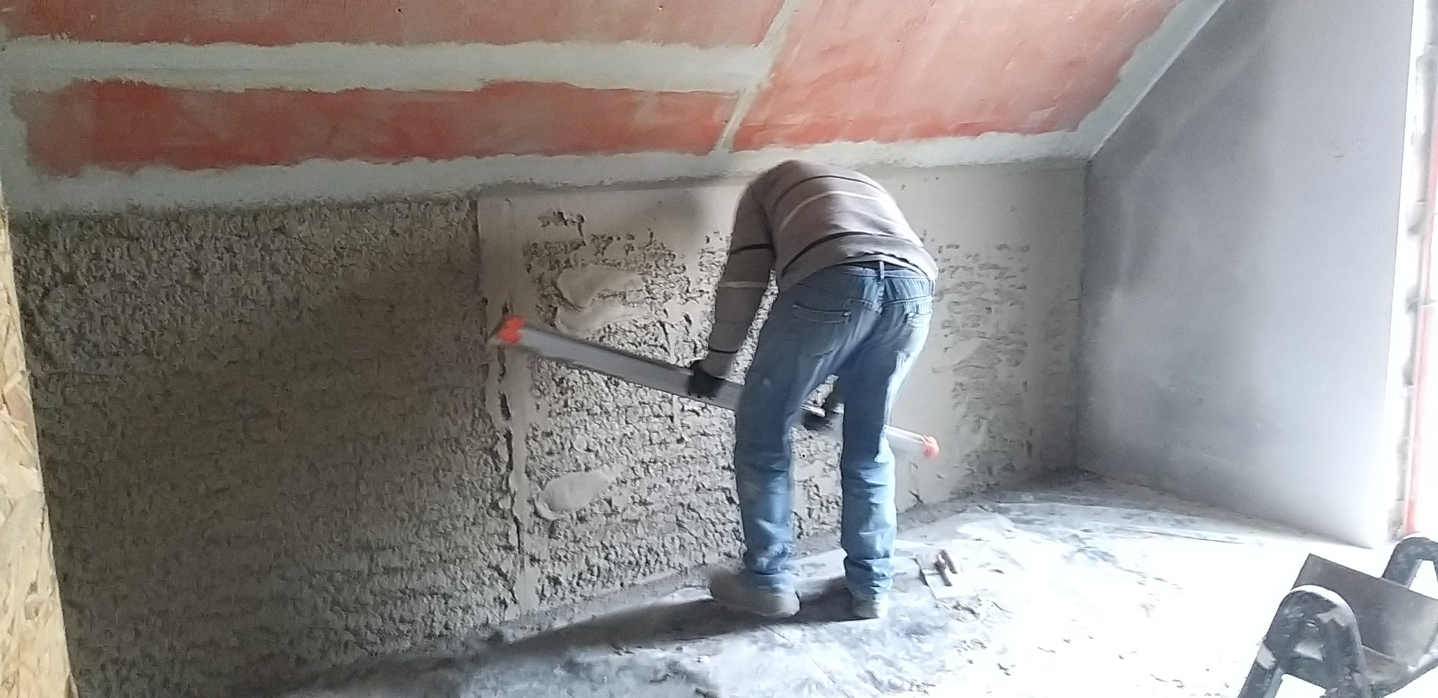 Praca tynkarska w poddaszu: mężczyzna w jeansach i swetrze wyrównuje szpachlą ścianę z surowym tynkiem. Widoczne narzędzia i fragment otynkowanego sufitu.