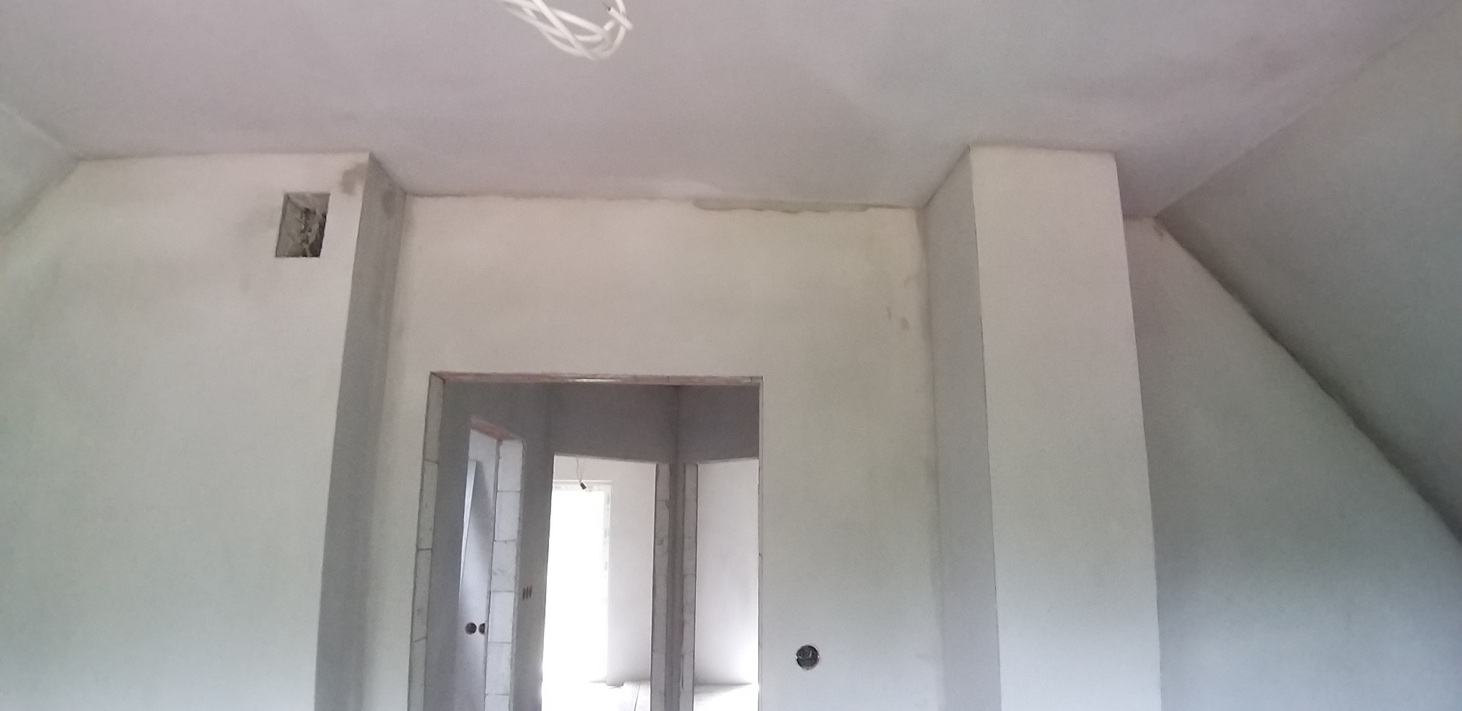 Wnętrze poddasza po tynkowaniu maszynowym. Widoczne gładkie ściany, otwory drzwiowe i fragment instalacji elektrycznej pod sufitem. Ściany w jasnym, szarym kolorze.