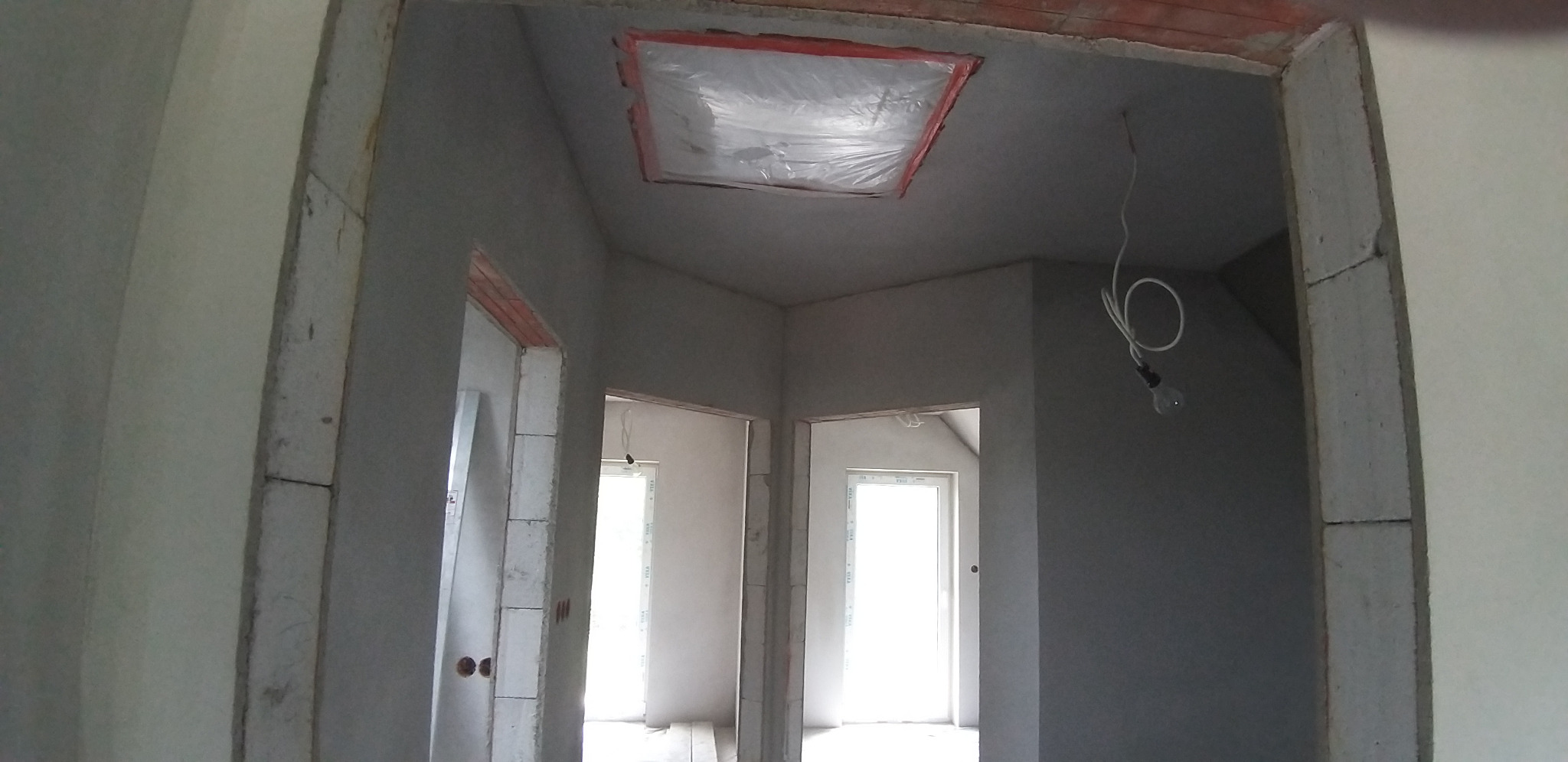 Wnętrze w trakcie tynkowania maszynowego: surowe ściany, widoczne otwory drzwiowe, zabezpieczony otwór w suficie folią. W tle okna wpuszczające jasne światło.