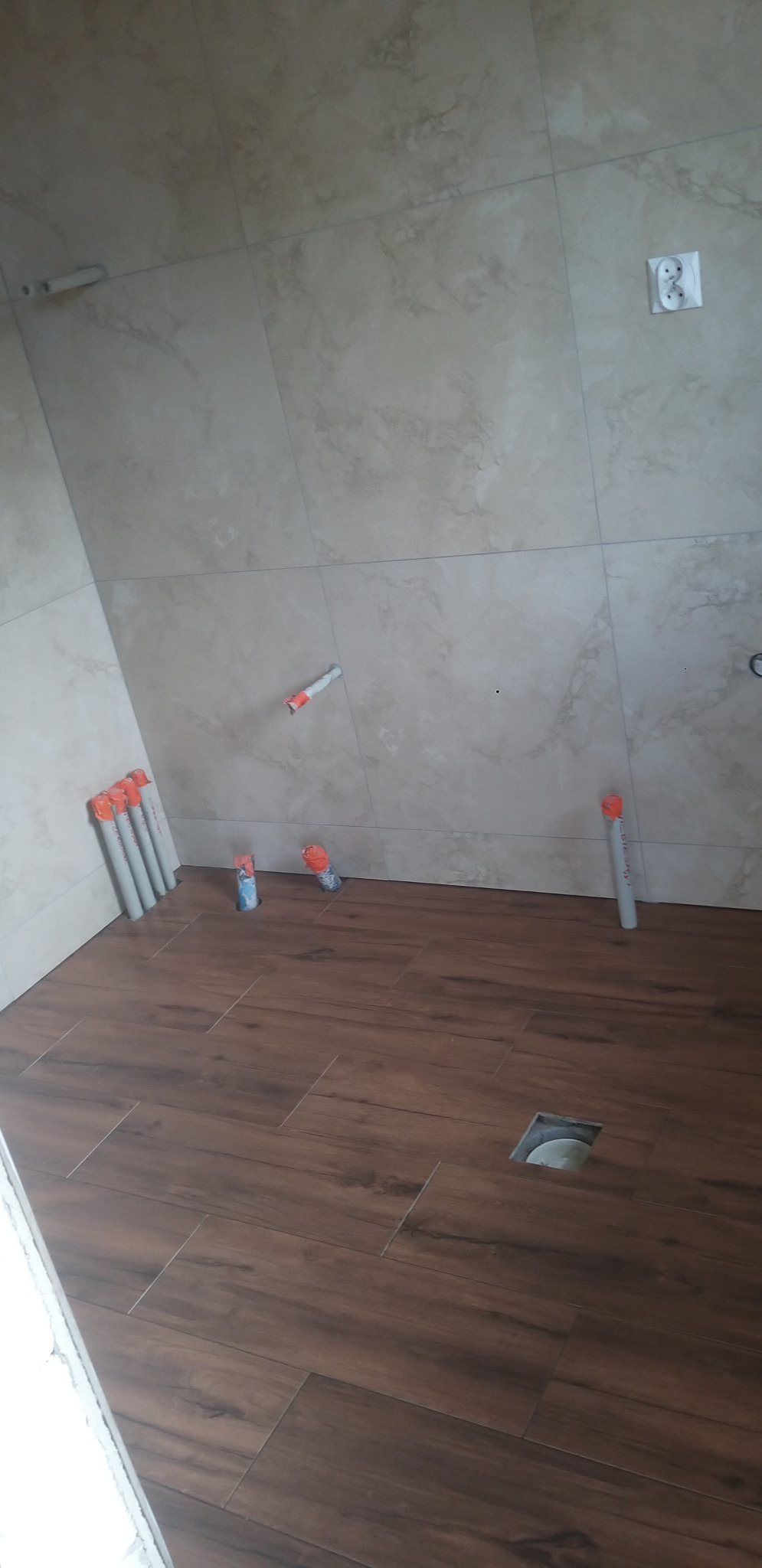 Wnętrze łazienki w trakcie remontu: brązowe płytki podłogowe imitujące drewno, beżowe płytki ścienne, rury kanalizacyjne wystające z podłogi i ściany, gniazdko elektryczne.