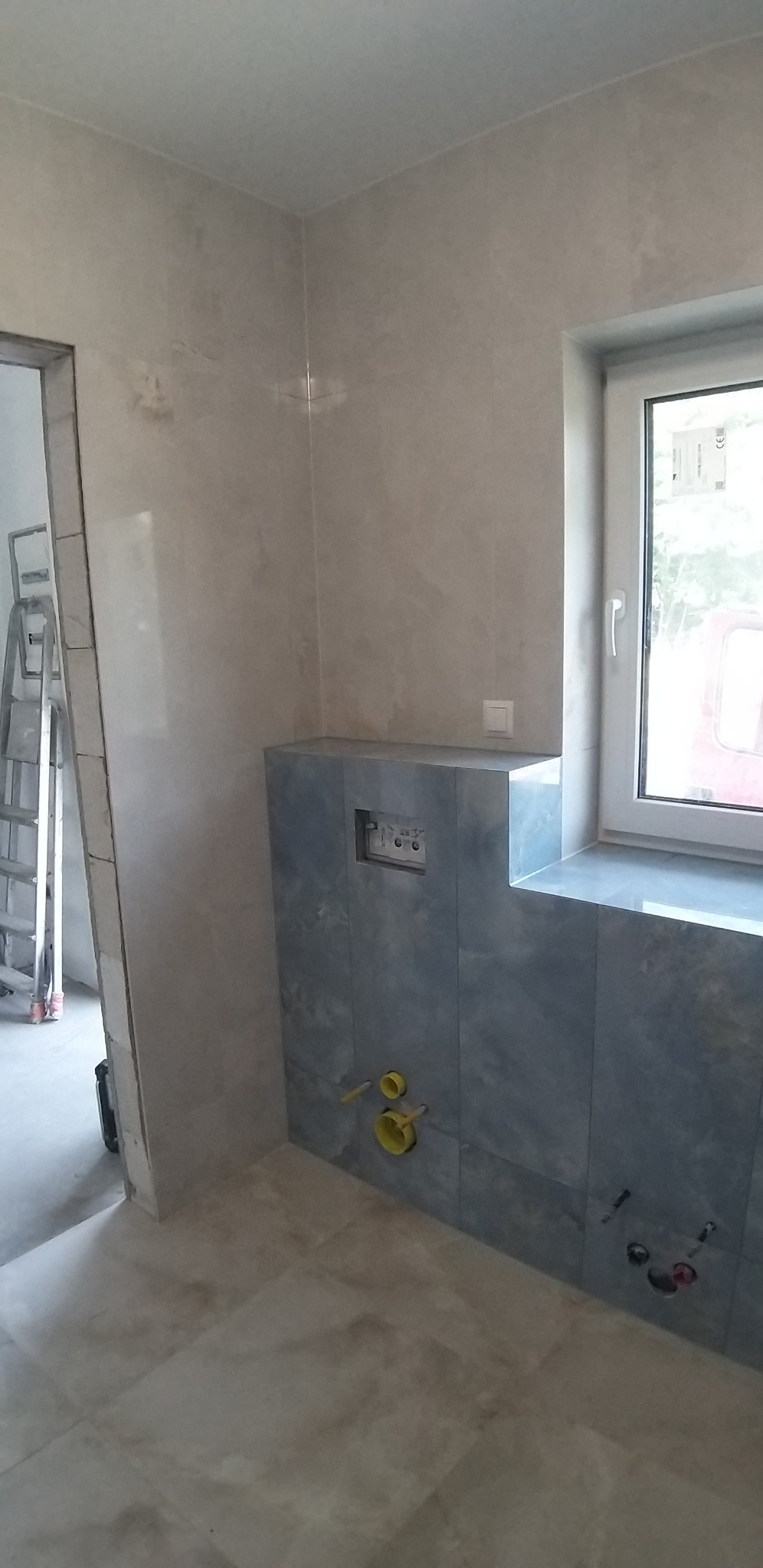 Wykończona łazienka z szaro-beżowymi płytkami na ścianach i podłodze. Widoczna zabudowa WC z niebieskimi płytkami i okno. W tle drabina malarska.