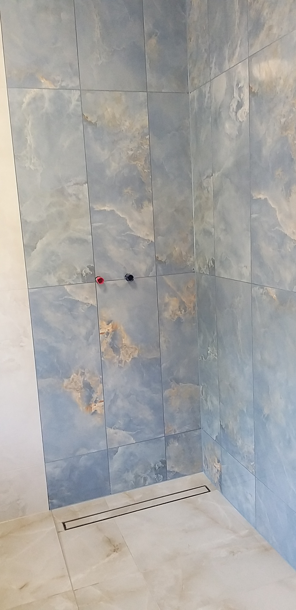 Wykończona łazienka z niebieskimi płytkami imitującymi marmur na ścianach i beżowymi na podłodze. Widoczny odpływ liniowy i przyłącza wodne.