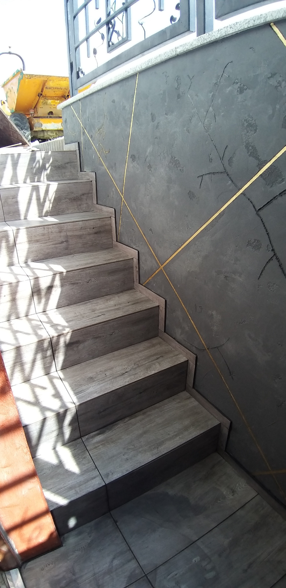Schody wyłożone płytkami imitującymi drewno, szara ściana z dekoracyjnymi złotymi liniami. W tle widoczny sprzęt budowlany. Ujęcie z dołu, wpadające światło.