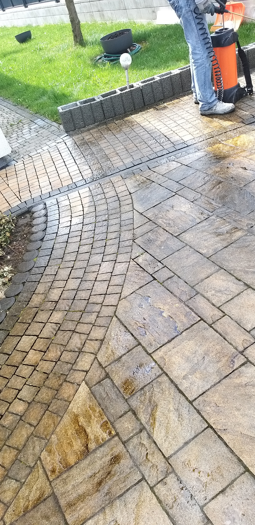 Czyszczenie kostki brukowej z użyciem myjki ciśnieniowej. Widoczne mokre i czyste fragmenty kostki, co sugeruje etap pracy w toku. Tło: trawnik i elementy dekoracyjne.