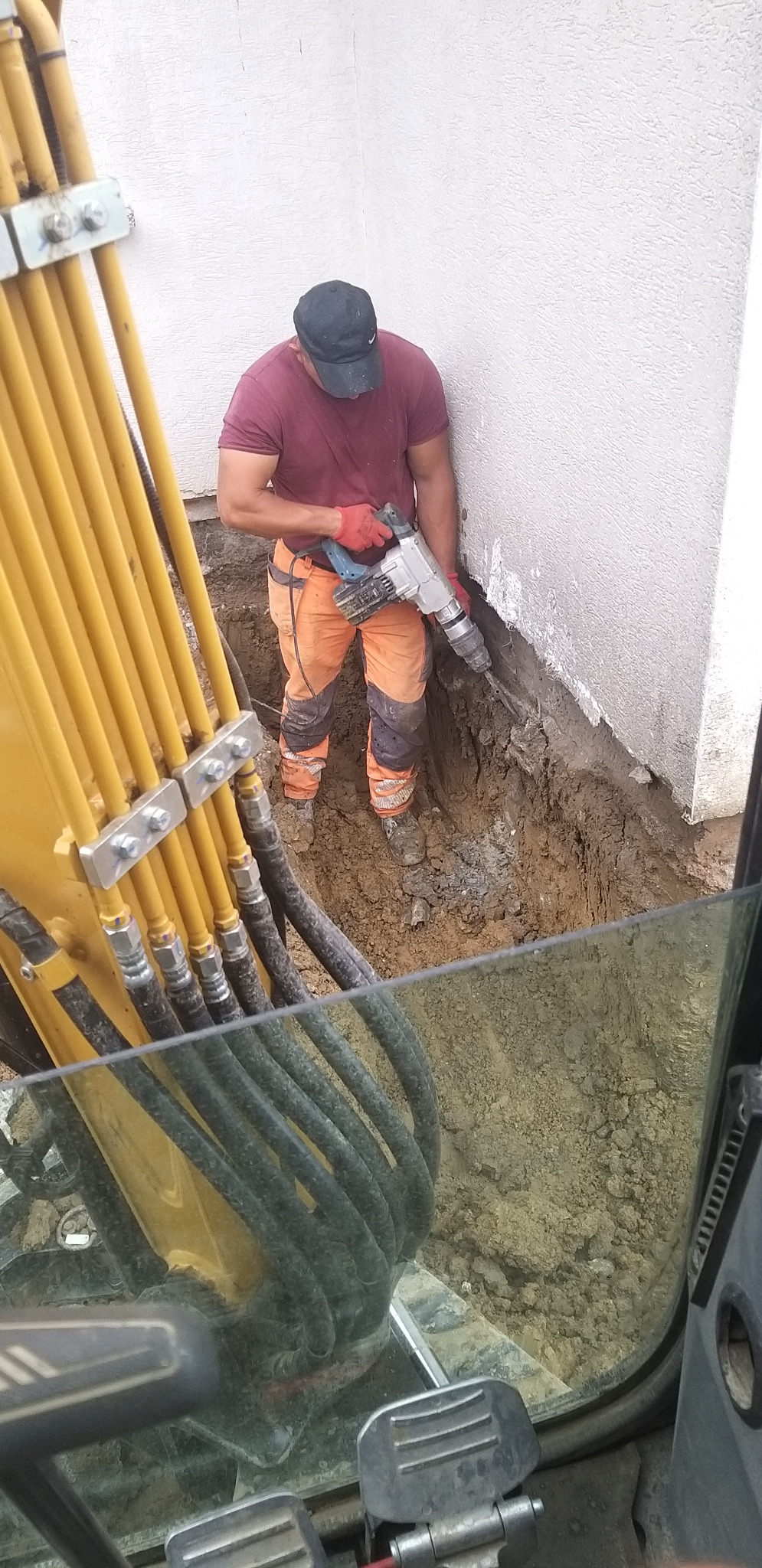 Pracownik w kasku i roboczych spodniach używa młota pneumatycznego przy fundamencie budynku. Widoczny fragment koparki z żółtymi przewodami.
