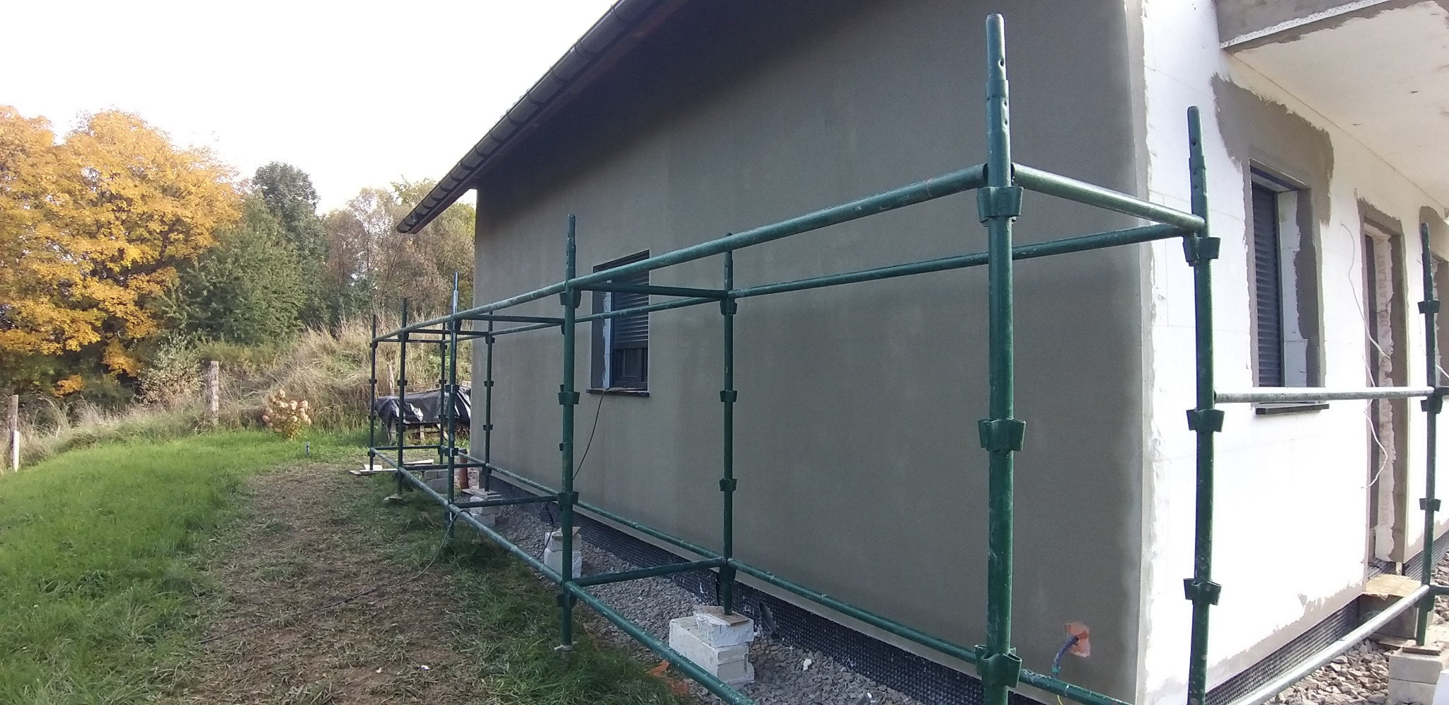 Ściana domu po tynkowaniu, z widocznymi rusztowaniami i oknem z roletą. Część elewacji w trakcie prac wykończeniowych. Jesienne drzewa w tle.