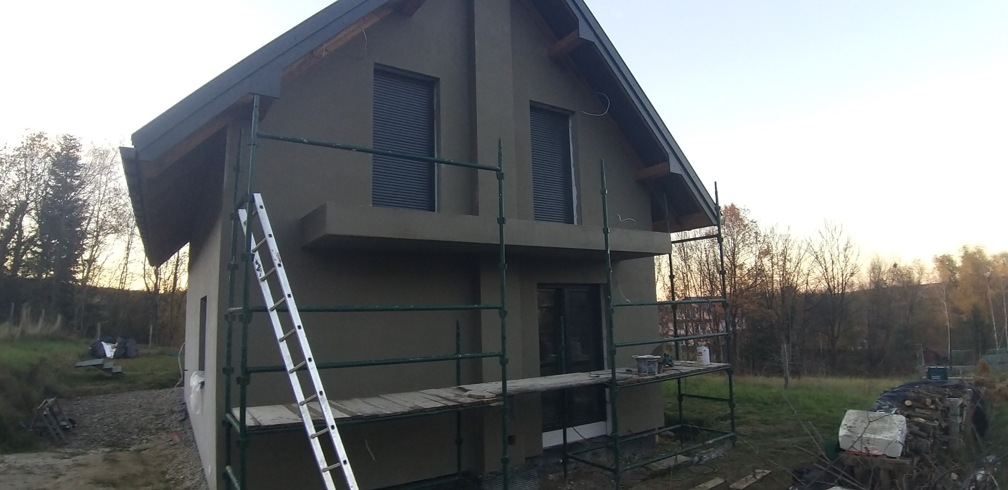 Dom jednorodzinny w trakcie tynkowania, widoczne rusztowania i drabina. Świeżo nałożony tynk w odcieniach szarości. Okna z roletami zewnętrznymi.