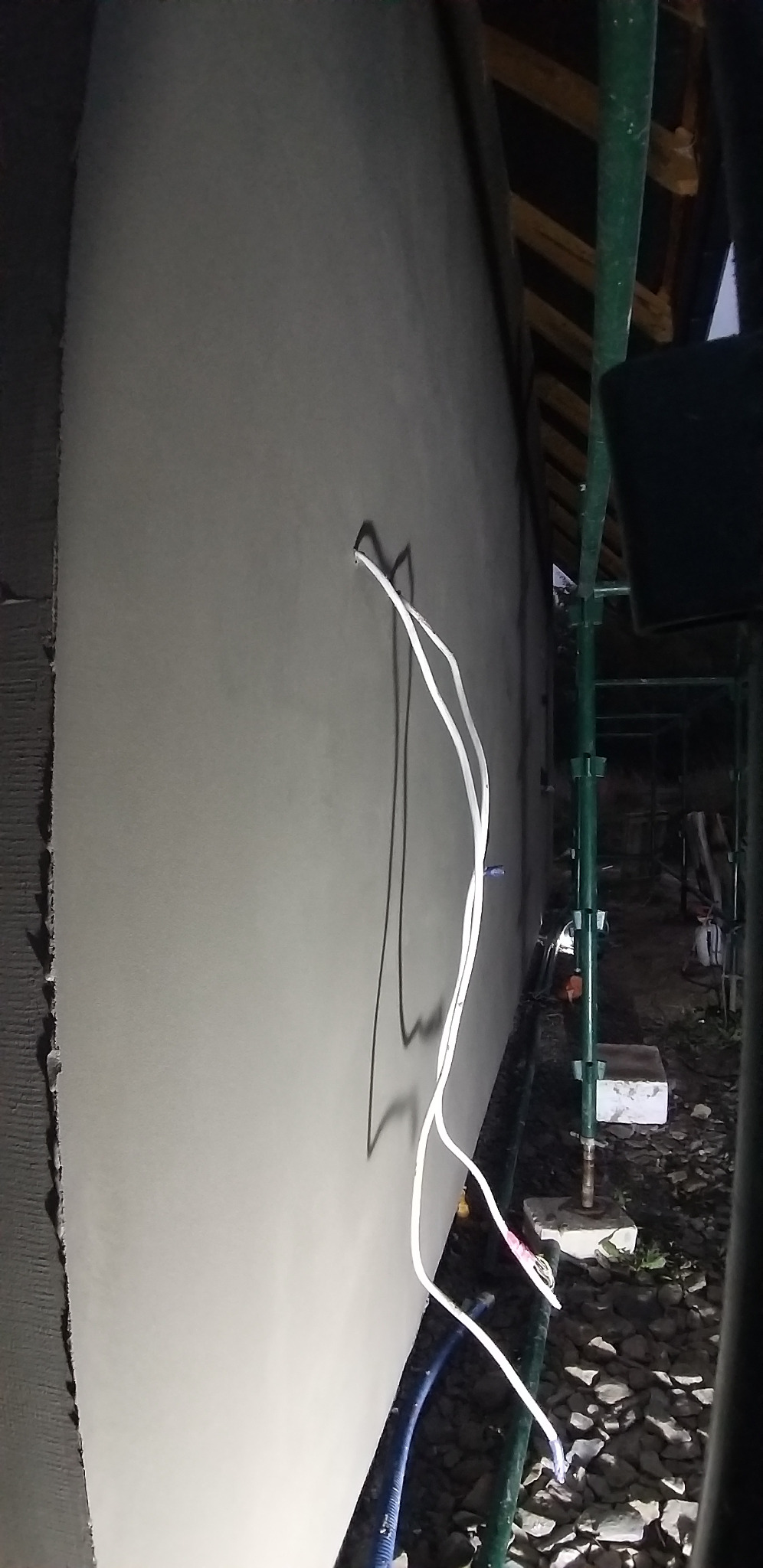 Świeżo otynkowana ściana zewnętrzna domu z wystającymi przewodami elektrycznymi, widoczne rusztowanie i fragment dachu. Nocne ujęcie.