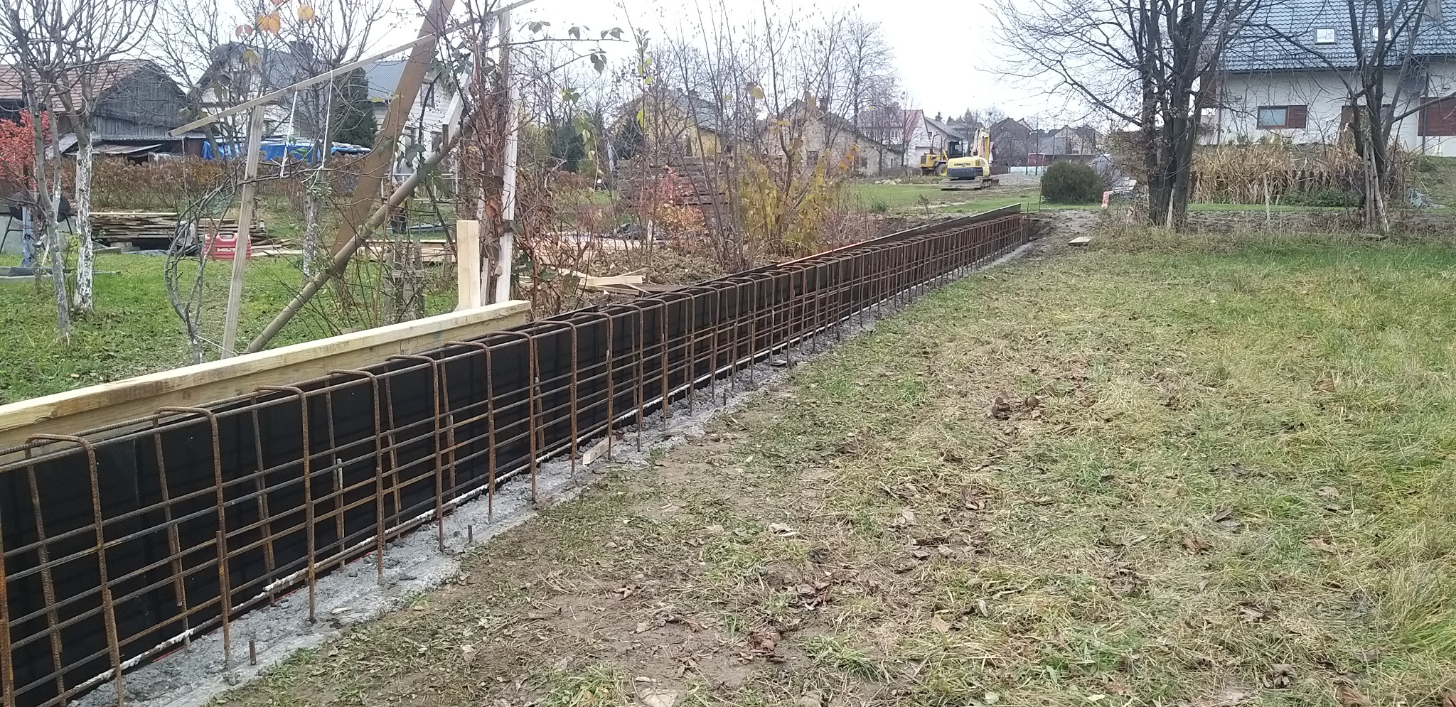 Zbrojenie fundamentu pod ogrodzenie z izolacją pionową, widoczny szalunek i wykop. W tle domy i koparka.