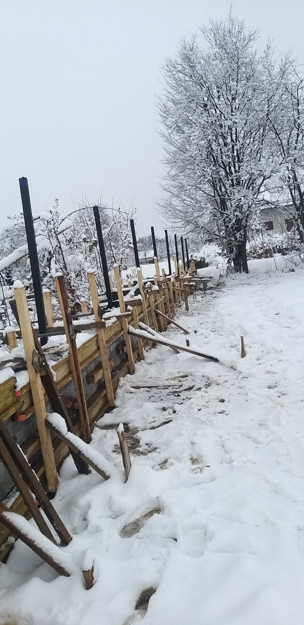 Zimowy krajobraz z widokiem na szalunek fundamentów w trakcie budowy. Śnieg pokrywa drewniane elementy i otaczający teren. W tle drzewa i budynek.