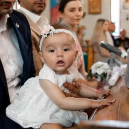 Urocza dziewczynka w białej sukience i opasce z kwiatami, siedząca na kolanach mężczyzny podczas uroczystości w kościele, z drewnianą ławką i innymi osobami w tle.