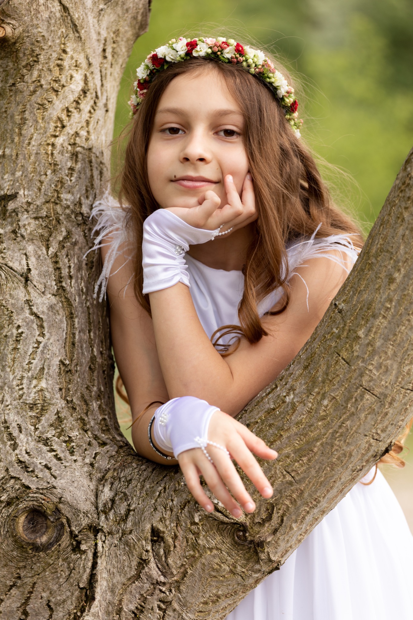 Dziewczynka w białej sukience i wianku z kwiatów opiera się o gałąź drzewa, spoglądając w obiektyw.