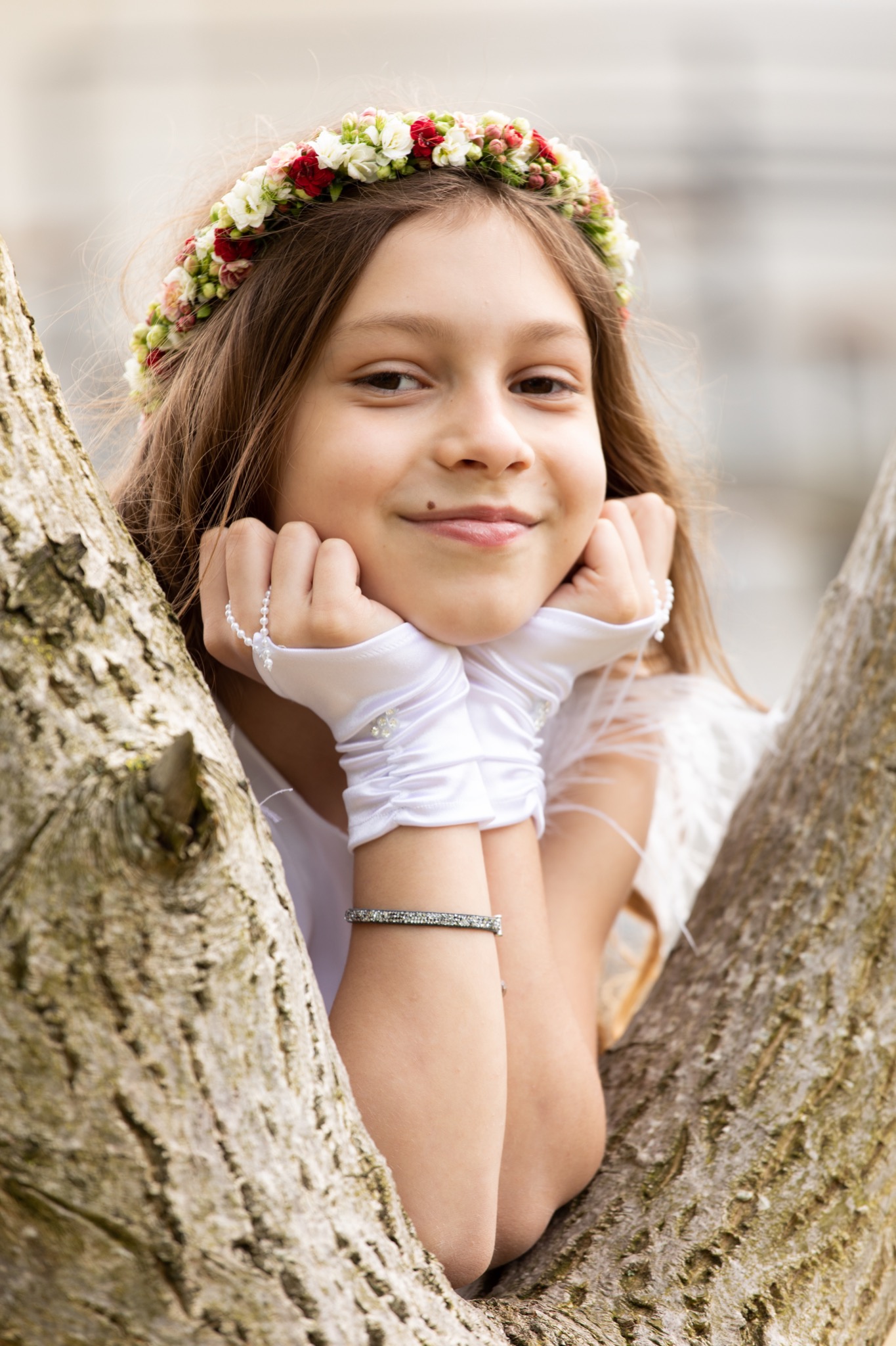 Uśmiechnięta dziewczynka w białej sukience i wianku z kwiatów, opierająca brodę na dłoniach w białych rękawiczkach, pozująca przy pniu drzewa.
