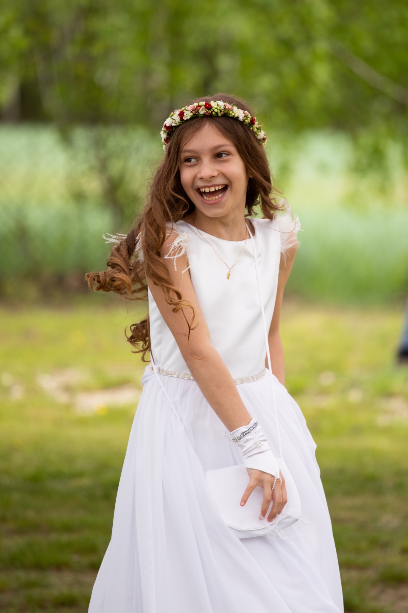 Uśmiechnięta dziewczynka w białej sukience komunijnej i wianku z kwiatów na głowie, trzymająca białą torebkę, na tle zieleni.