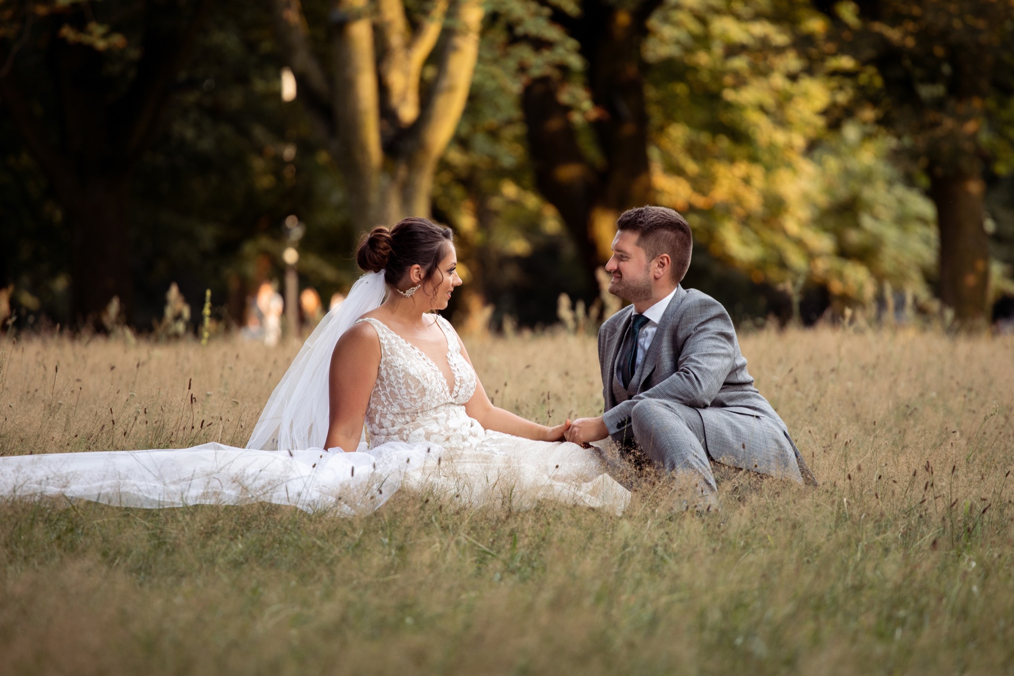Para młoda siedząca na trawie, panna młoda w koronkowej sukni z trenem i welonem, pan młody w szarej kraciastej marynarce, oboje trzymają się za ręce i patrzą na siebie z uśmiechem, w tle drzewa.