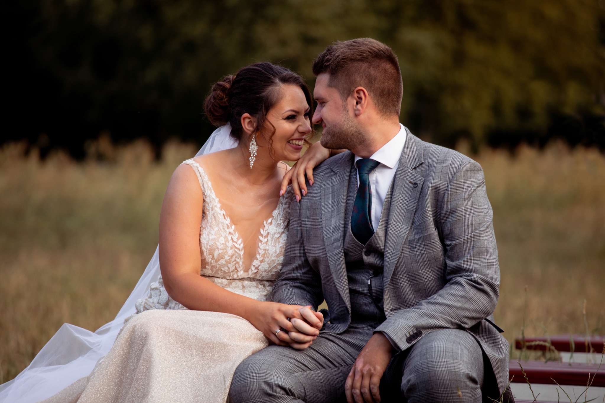 Uśmiechnięta para młoda siedzi blisko na ławce na tle łąki, panna młoda w sukni z koronkową górą i długimi kolczykami, pan młody w szarym garniturze w kratę i ciemnozielonym krawacie.