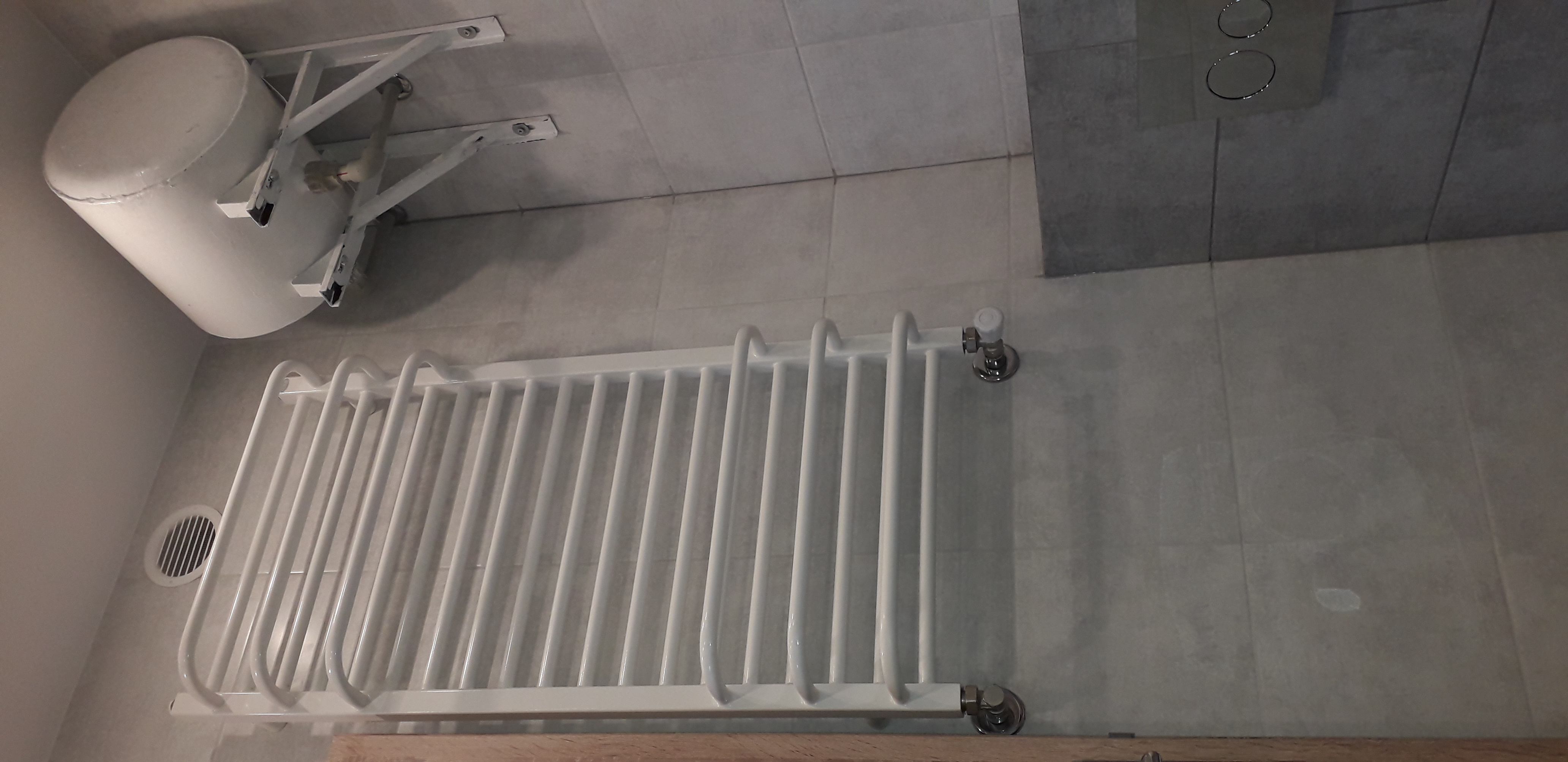 Biały grzejnik łazienkowy drabinkowy, zamontowany na szarej, kafelkowanej ścianie, widoczny zbiornik wyrównawczy i panel sterowania spłuczką.