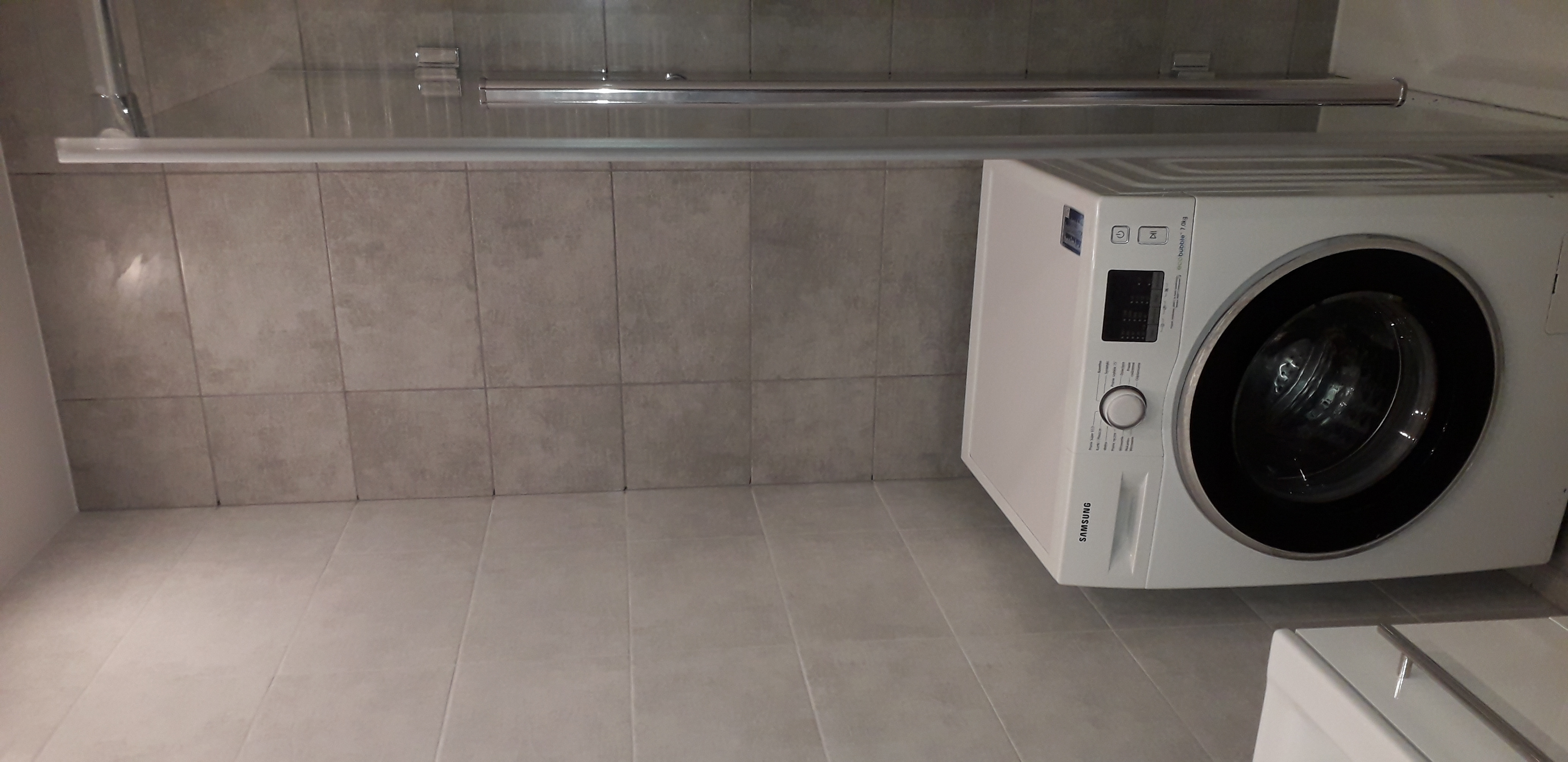 Nowoczesna łazienka z pralką Samsung Eco Bubble 7.0 kg i szarymi płytkami ceramicznymi na ścianach i podłodze, widoczna kabina prysznicowa.