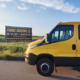 Żółty samochód pomocy drogowej zaparkowany na poboczu drogi, w tle tablica informacyjna z numerem telefonu i adresem strony internetowej.