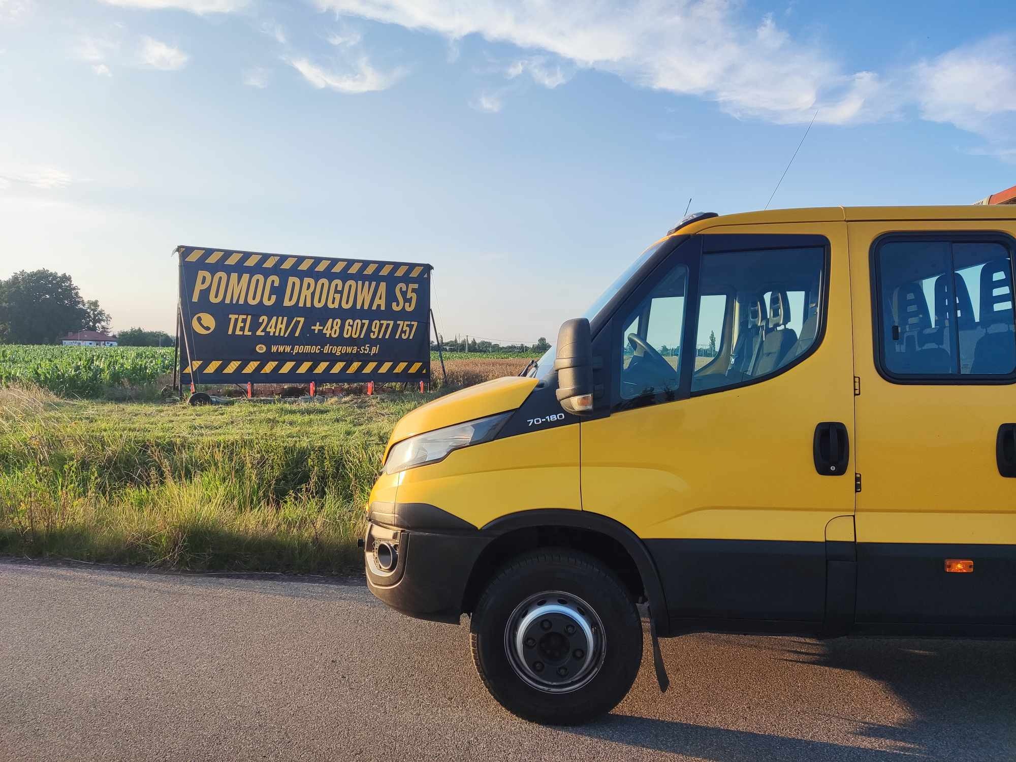 Żółty samochód pomocy drogowej zaparkowany na poboczu drogi, w tle tablica informacyjna z numerem telefonu i adresem strony internetowej.
