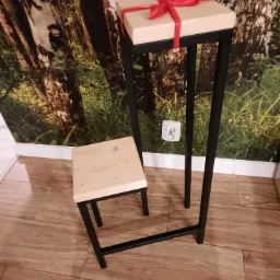 Metalowy kwietnik z drewnianym blatem, na kt&oacute;rym leży prezent z czerwoną wstążką, obok mały drewniany stołek na tle fototapety z motywem leśnym.