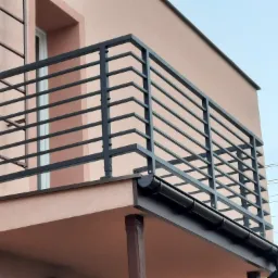 Czarna, pozioma balustrada balkonowa z prostokątnymi poprzeczkami, zamontowana na elewacji budynku o jasnoróżowym odcieniu. Widoczna rynna odprowadzająca wodę.