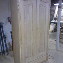 Krystian Dosz - Niedokończona, dwudrzwiowa szafa z jasnego drewna, widoczne detale wykończeniowe, surowe wnętrze warsztatu w tle.