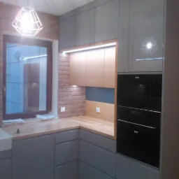 Szara kuchnia z ceglaną ścianą i drewnianym blatem. Widoczne górne szafki, piekarnik w zabudowie i lampa wisząca nad oknem. Nowoczesny styl.