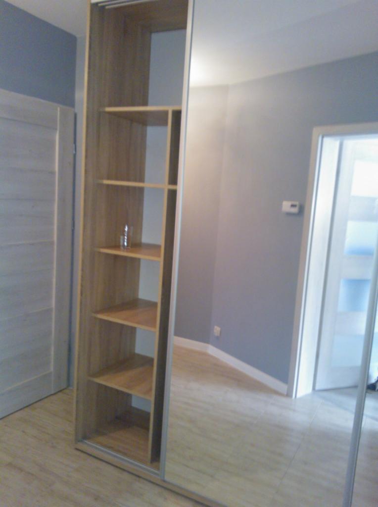 Wysoki regał z jasnego drewna z otwartymi półkami, wbudowany w szafę z lustrzanymi drzwiami przesuwnymi, odbijający wnętrze pokoju z szarymi ścianami i drewnianą podłogą.