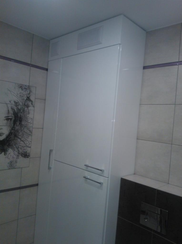 Wysoka, biała szafka łazienkowa z dwoma frontami i chromowanymi uchwytami, dopasowana do sufitu, umieszczona w rogu łazienki z płytkami w odcieniach szarości i czarnym akcentem.