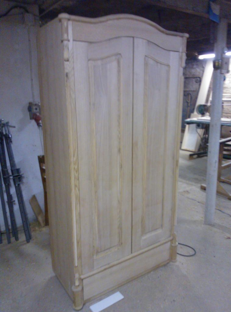 Niedokończona, dwudrzwiowa szafa z jasnego drewna, widoczne detale wykończeniowe, surowe wnętrze warsztatu w tle.