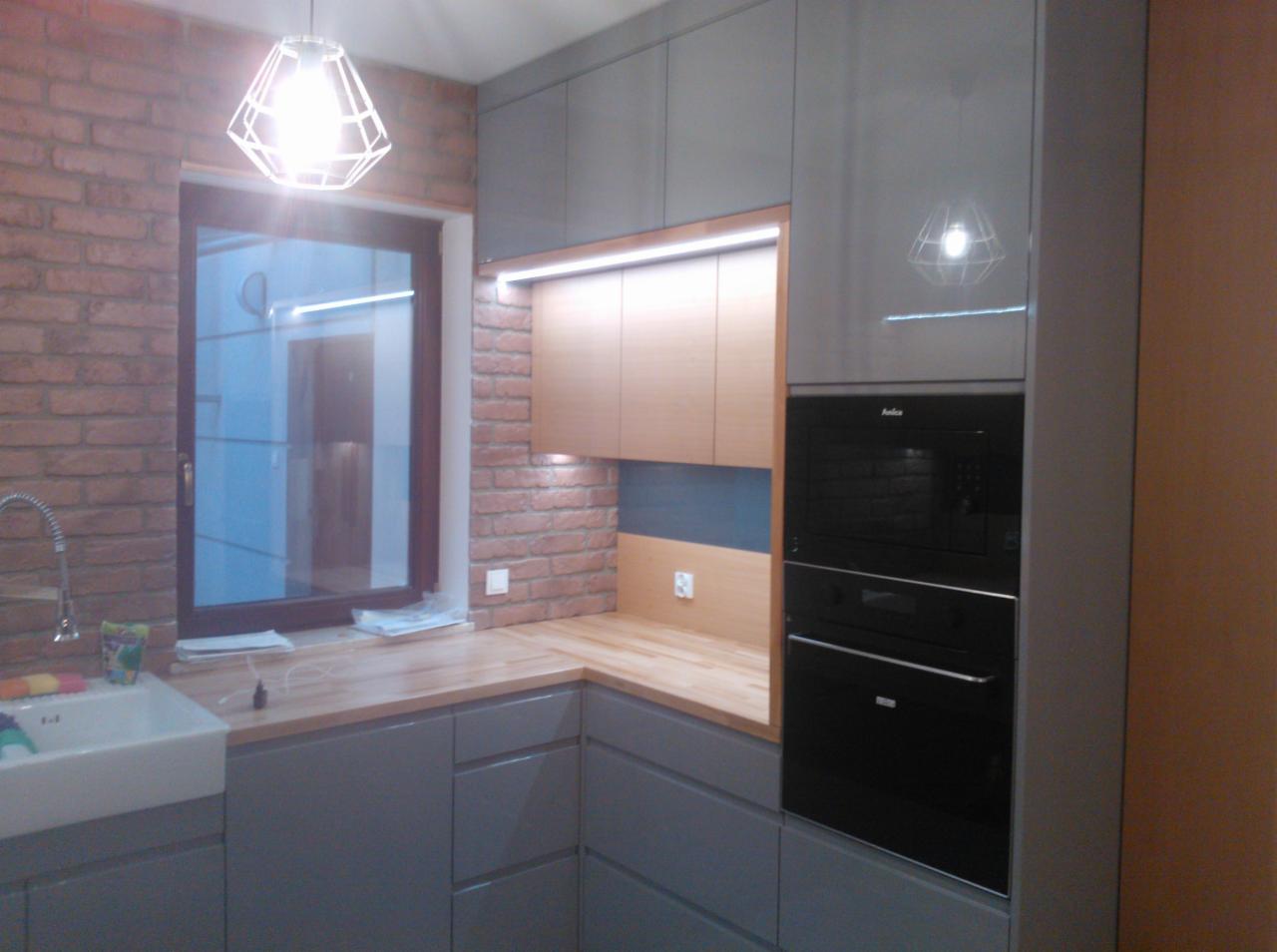 Szara kuchnia z ceglaną ścianą i drewnianym blatem. Widoczne górne szafki, piekarnik w zabudowie i lampa wisząca nad oknem. Nowoczesny styl.