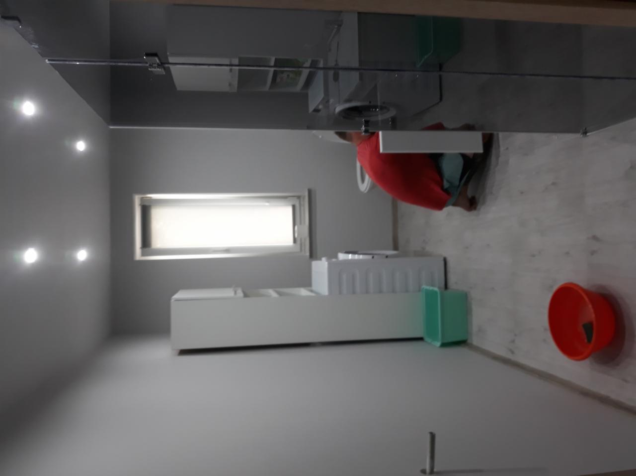 Wnętrze łazienki w trakcie prac wykończeniowych: widoczna biała zabudowa, okno z żaluzjami, osoba w czerwonej koszulce montująca toaletę, oraz plastikowe miski na podłodze.