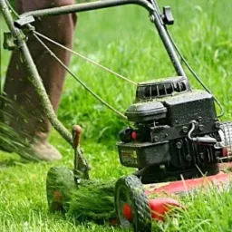 Kosiarka spalinowa w trakcie pracy, wyrzucająca skoszoną trawę, widoczne nogi operatora w tle.