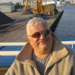 Mężczyzna w okularach na tle nabrzeża z dźwigiem i stertami materiałów. Ujęcie z dołu, ciepłe światło.