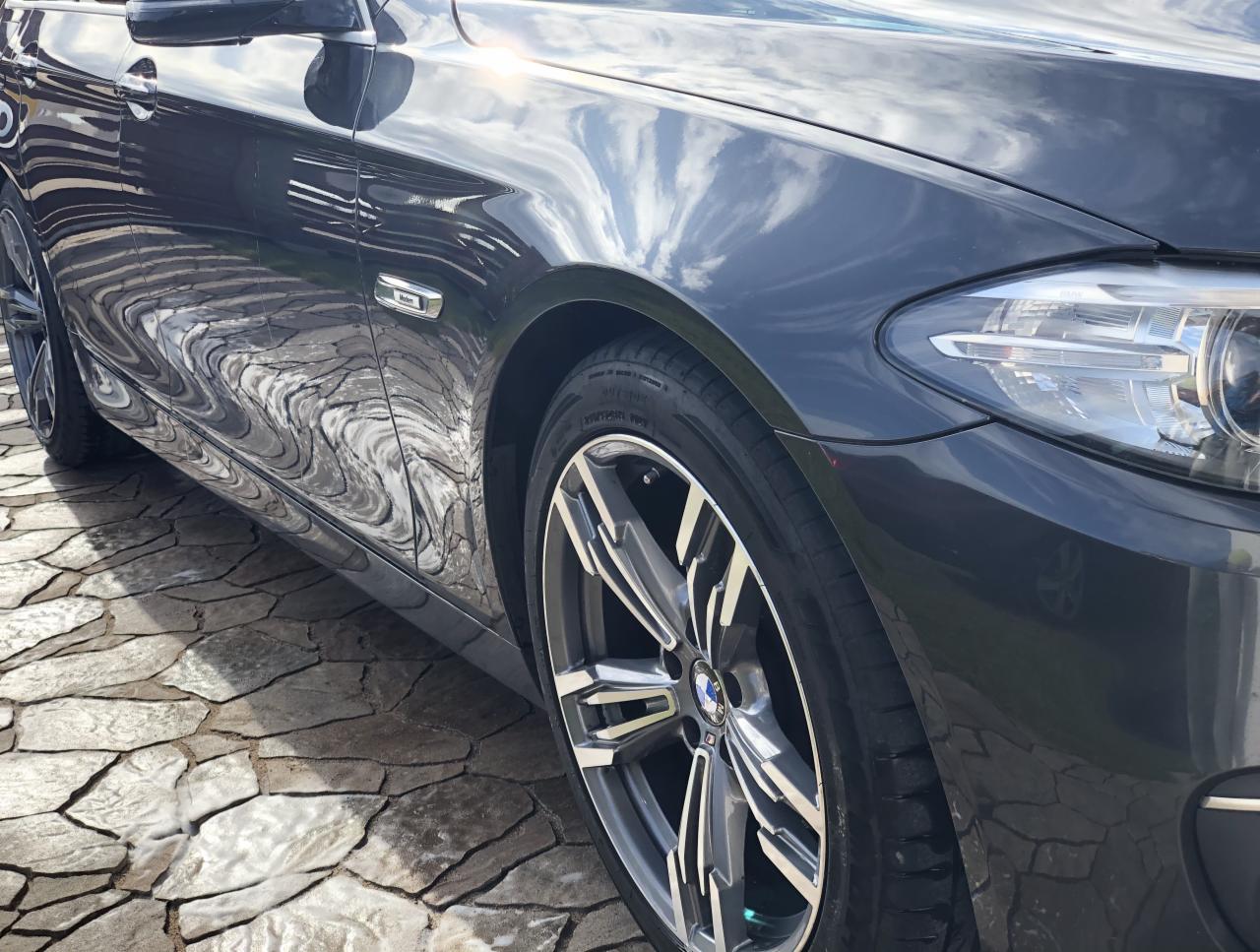 Błyszczący, grafitowy samochód BMW po umyciu, z widocznym odbiciem chmur na karoserii, stojący na wyczyszczonej kostce brukowej.