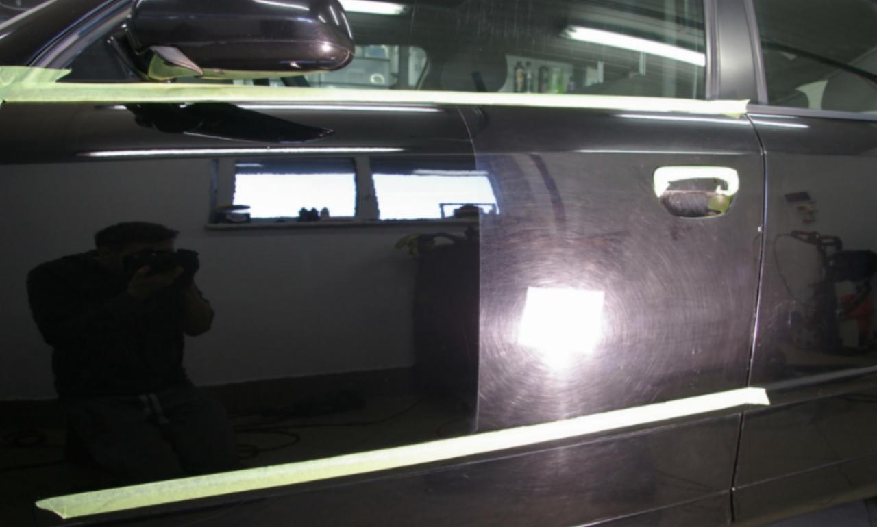 Częściowo wypolerowane, czarne drzwi samochodu z widocznymi śladami polerowania i odbiciem fotografa robiącego zdjęcie. Elementy karoserii zabezpieczone taśmą. Widoczne zarysowania przed polerowaniem.
